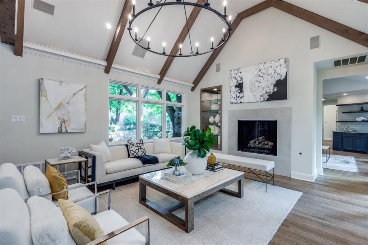 A living room with a vaulted ceiling, painting, fireplace, windows, white couch, white chairs, and a center table with a vase.