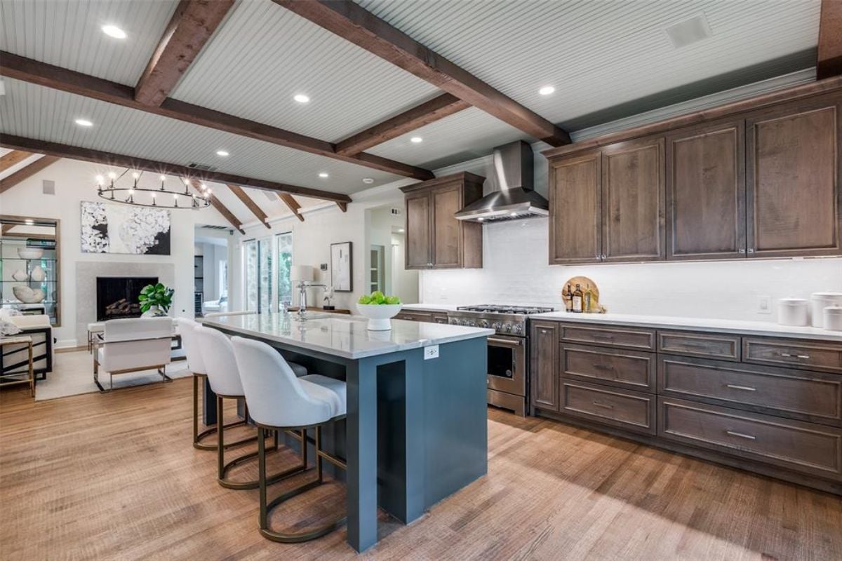 A kitchen with an exposed beam ceiling, wooden floor, island, stools, countertop, and cabinets.