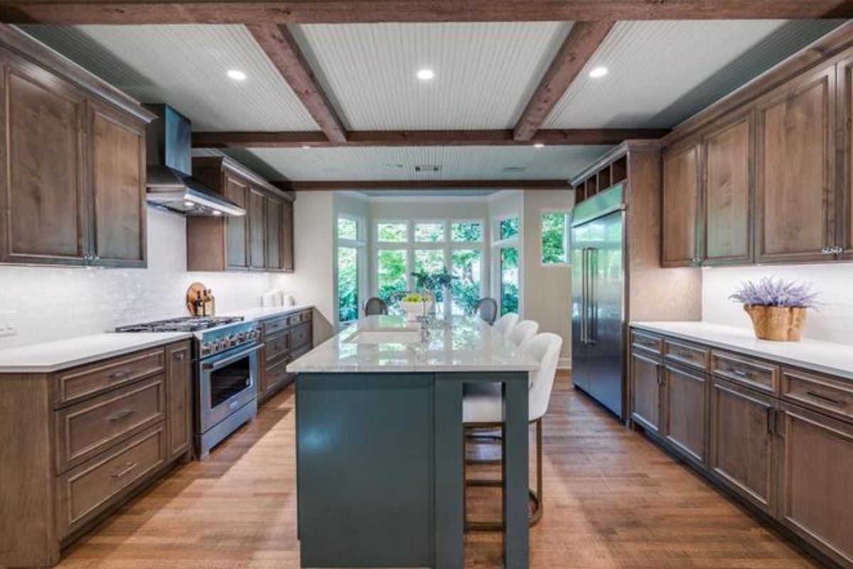 A kitchen with an exposed beam ceiling, wooden floor, island, stools, countertop, and cabinets.