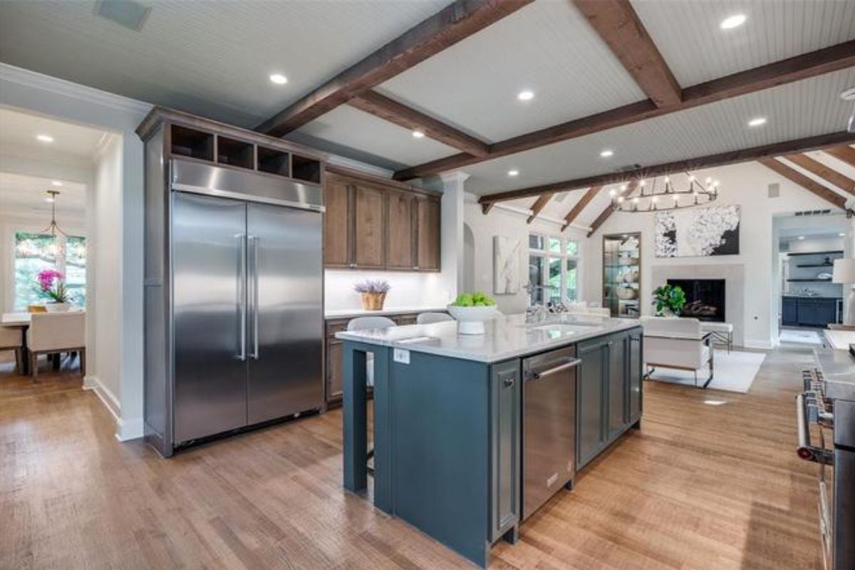 A kitchen with an exposed beam ceiling, wooden floor, island, stools, countertop, and cabinets.