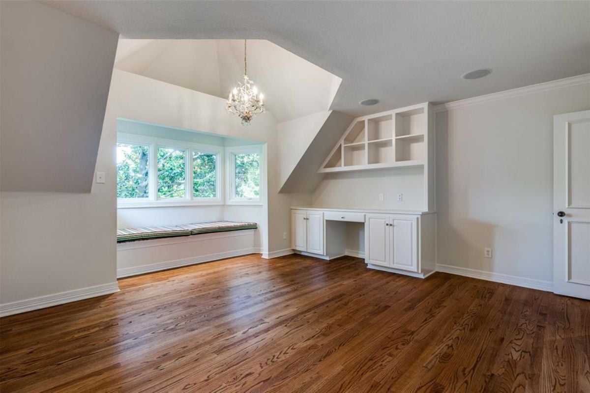 An empty room with a white ceiling, windows, chandelier, cabinets, and a wooden floor.