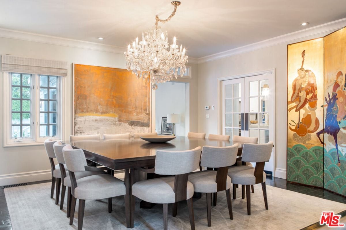 A dining room with paintings, a chandelier, windows, and a dining table surrounded by chairs.
