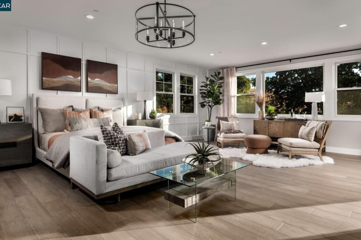 A bedroom with nightstands, windows, cabinets, paintings, a couch, chairs, and a coffee table.