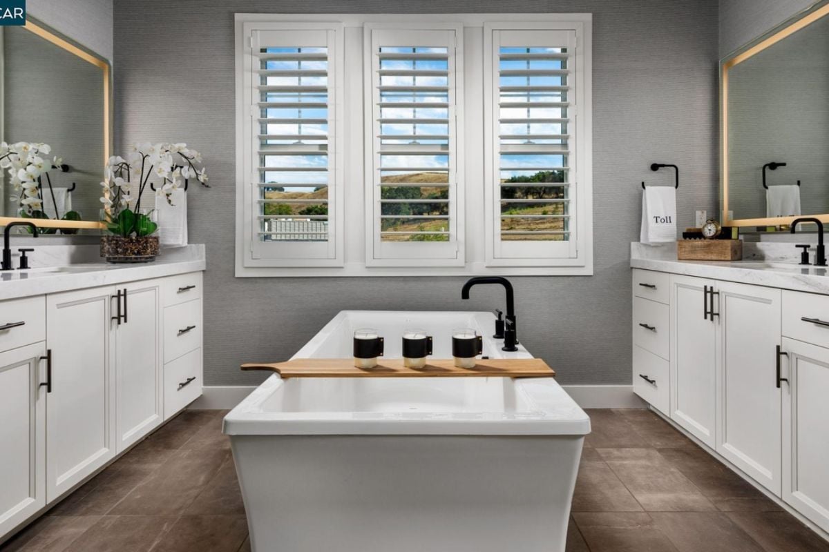 A bathroom with vanities, cabinets, mirror, window, and bathtub.
