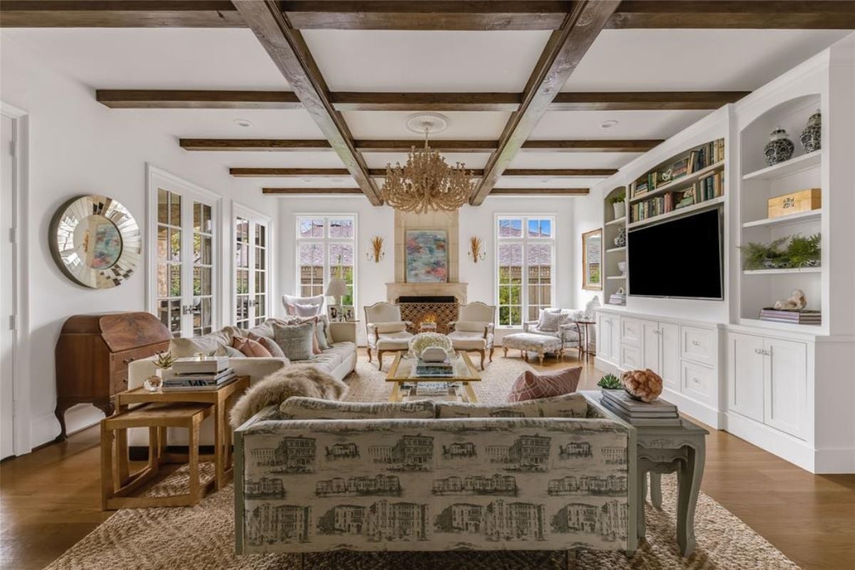 A living room with an exposed beam ceiling, chandelier, TV, shelves, cabinets, couch, chair, and center table.