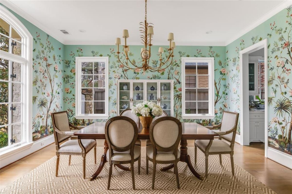 A dining room with wallpaper, chandelier, windows, and a dining table surrounded by chairs.
