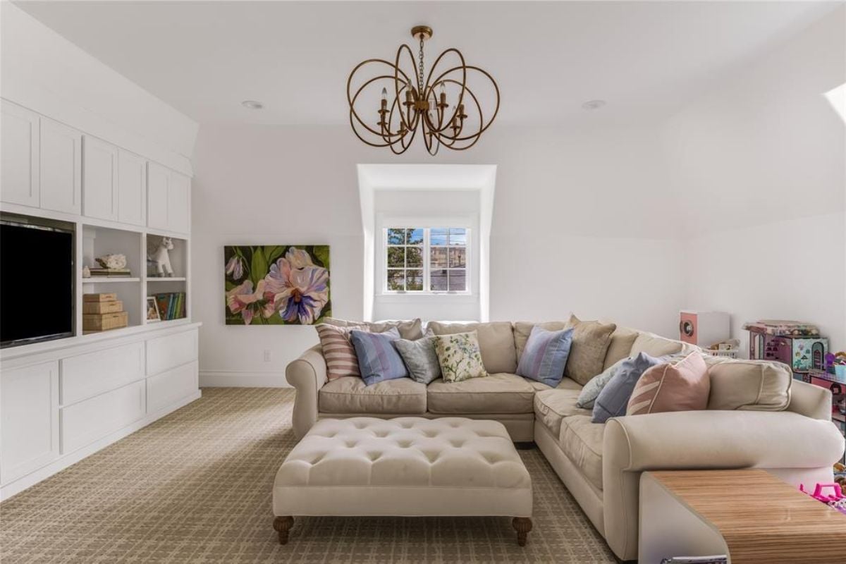 A lounge with a chandelier, window, TV, shelf, cabinets, couches, and an ottoman.