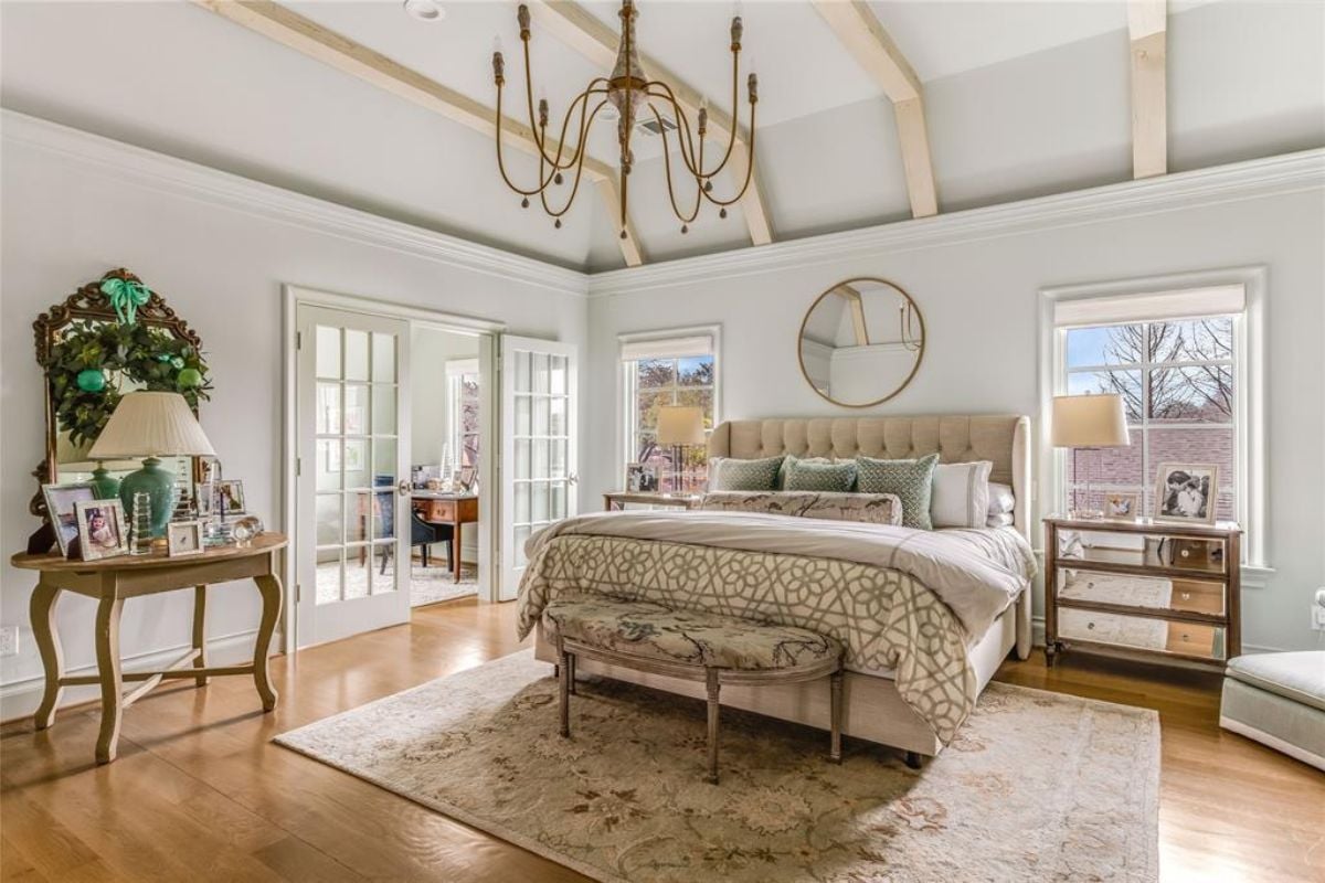 A bedroom with a nightstand, exposed beam ceiling, windows, rug, and table.