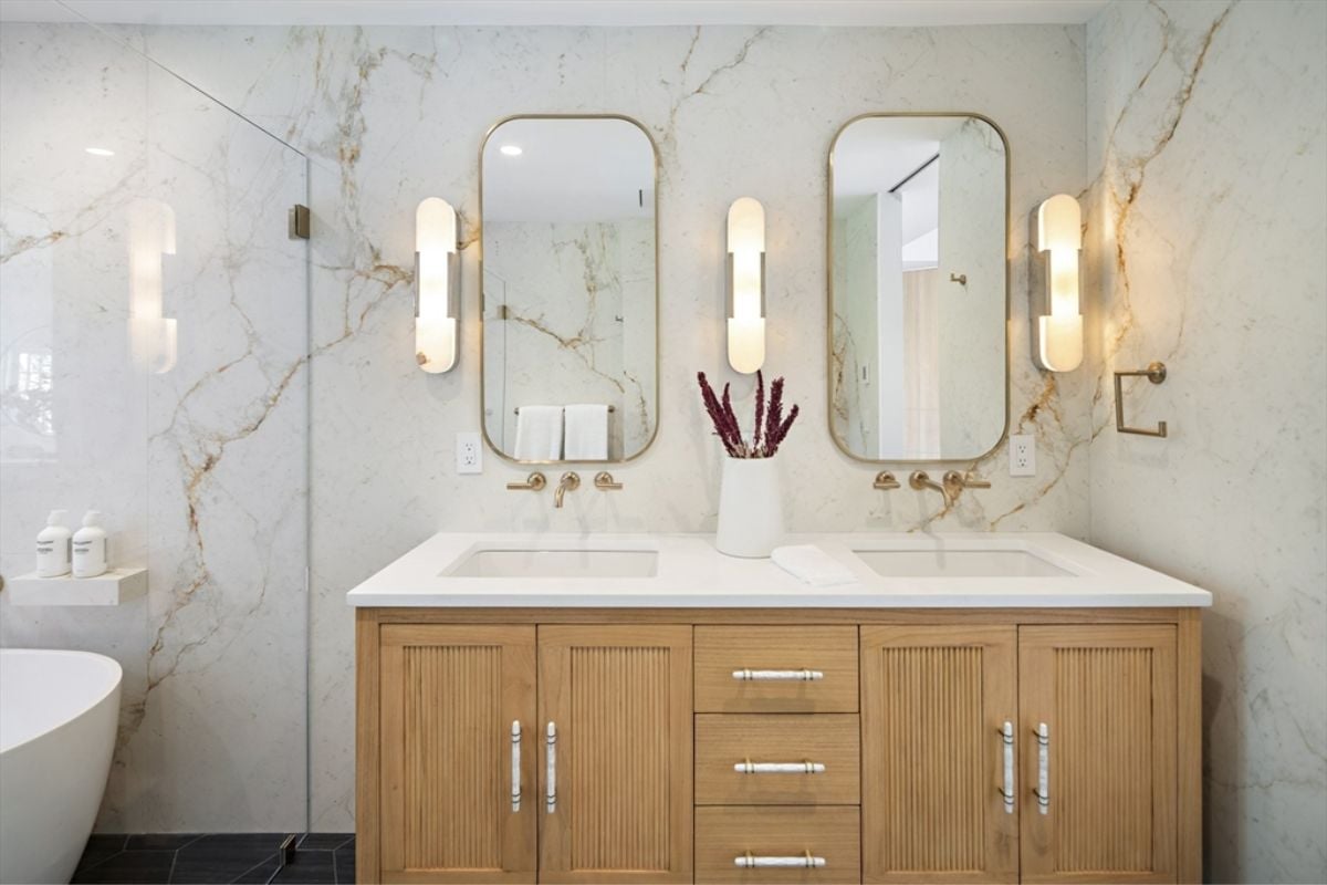 A bathroom with marble walls, mirrors, countertop, sinks, and cabinets.