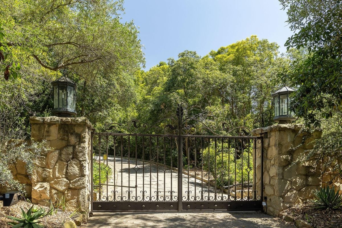 There is a front gate entry with a metal gate, plants, and trees.