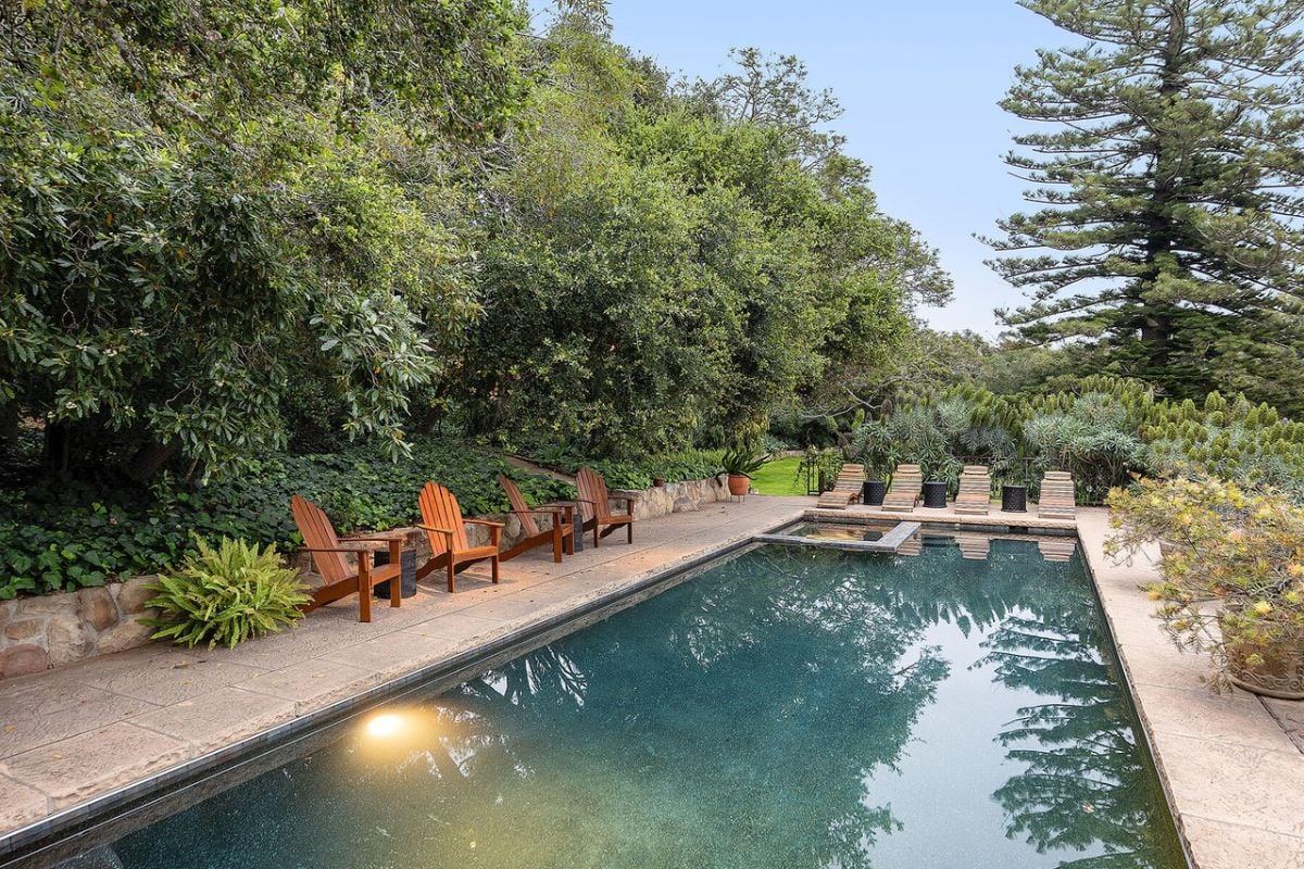 There is a swimming pool with chairs on the poolside, surrounded by plants and trees.