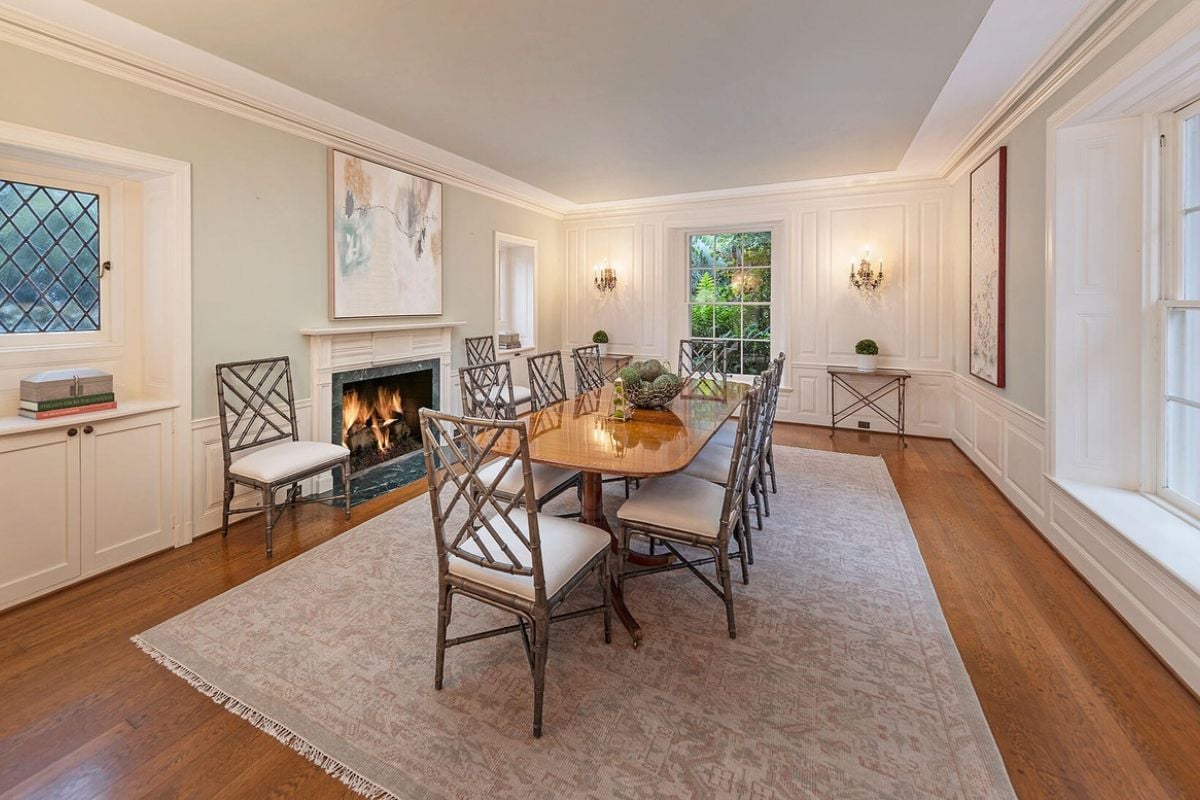 A dining room with a rug, fireplace, paintings, and a dining table surrounded by chairs.