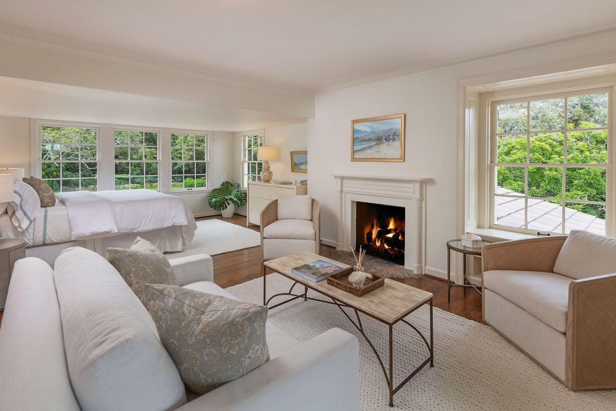 A bedroom with a bed, windows, cabinet, painting, fireplace, couches, and a center table.