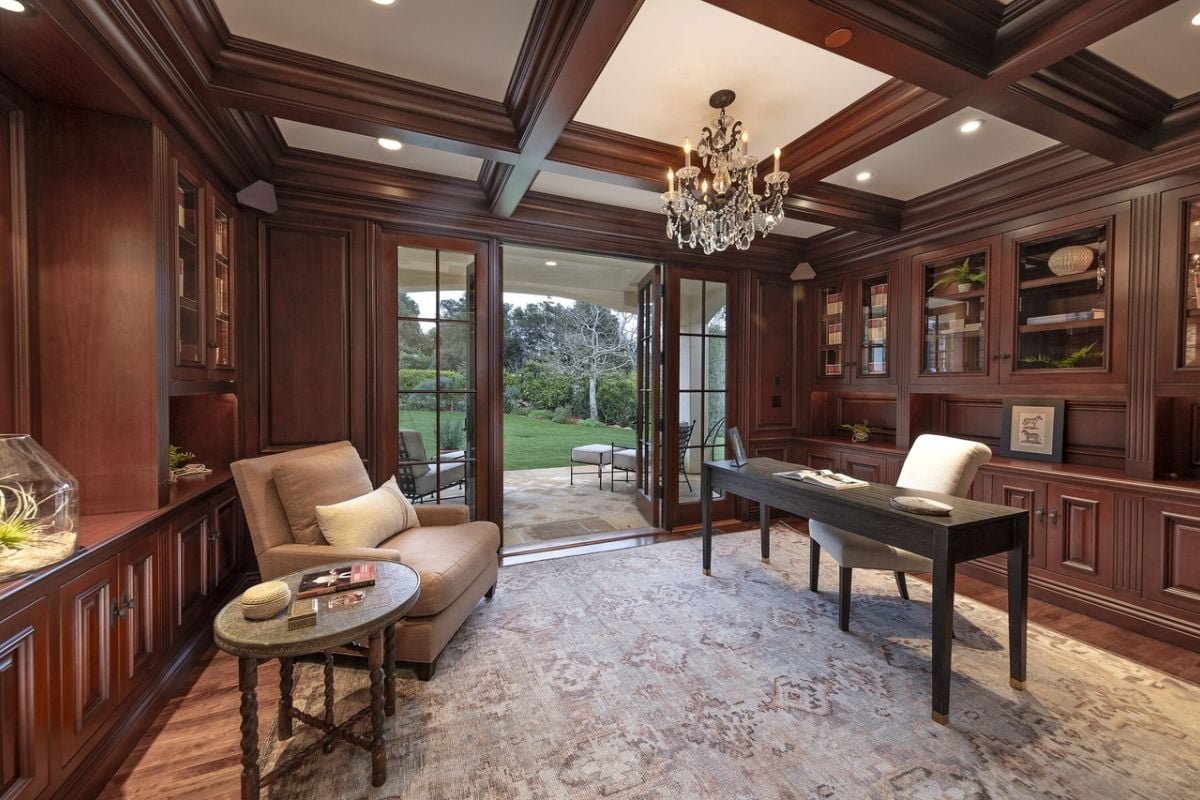 A home office with an exposed beam ceiling, chandelier, cabinets, a single couch, coffee table, desk, and chair.