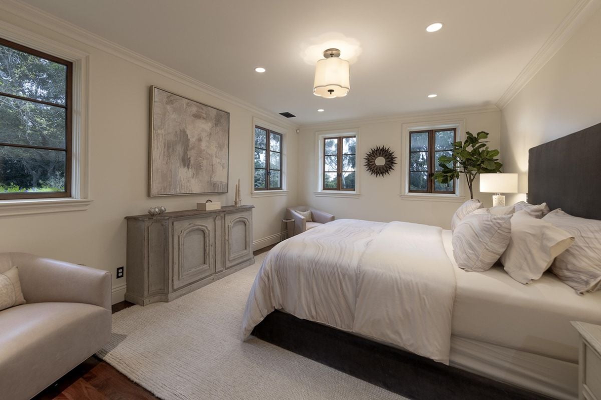 A bedroom with a bed, cabinet, painting, windows, a houseplant, and two studio couches.