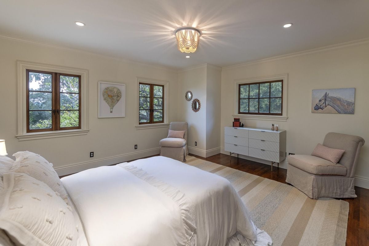 A bedroom with a bed, cabinet, painting, windows, and tchairs