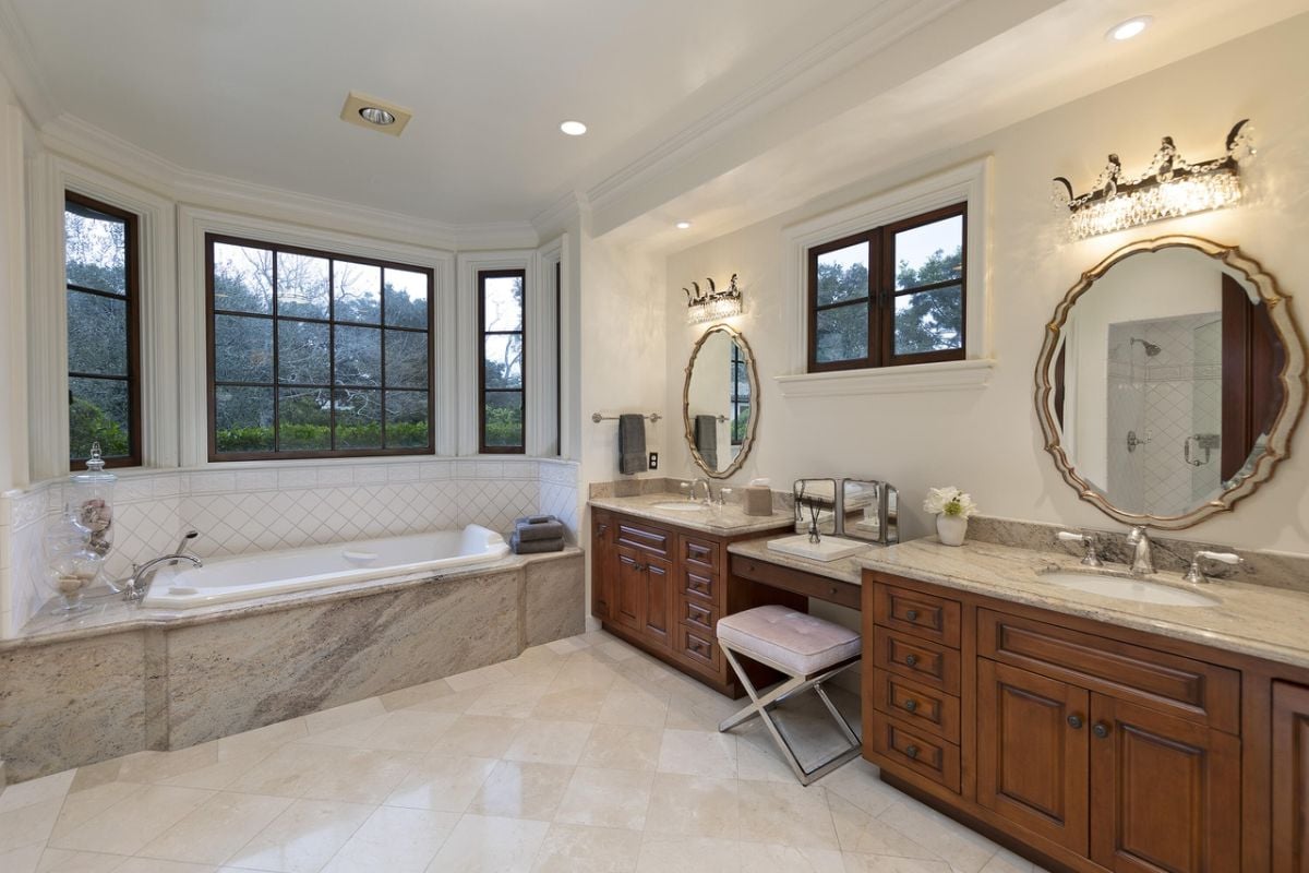 A bathroom with vanities, mirrors, a chair, windows, and a bathtub.
