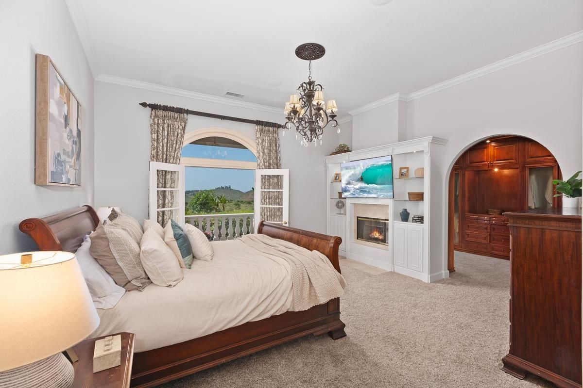 A bedroom with a bed, chandelier, painting, TV, fireplace, and cabinets.