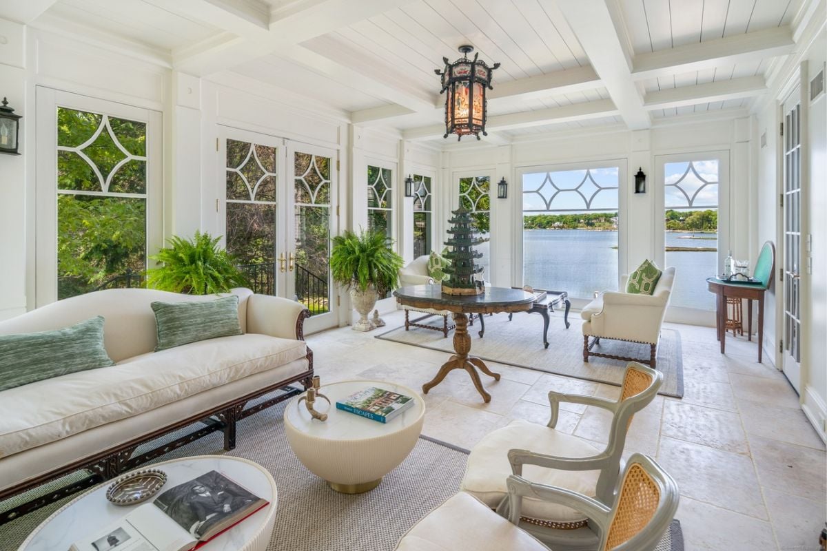 A living room with an exposed beam ceiling, windows, houseplants, a couch, chairs, and coffee tables.
