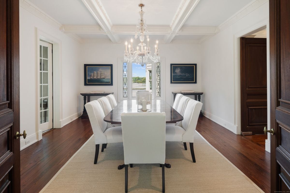 A dining room with a chandelier, white ceiling, paintings, a rug, and a dining table surrounded by white chairs.