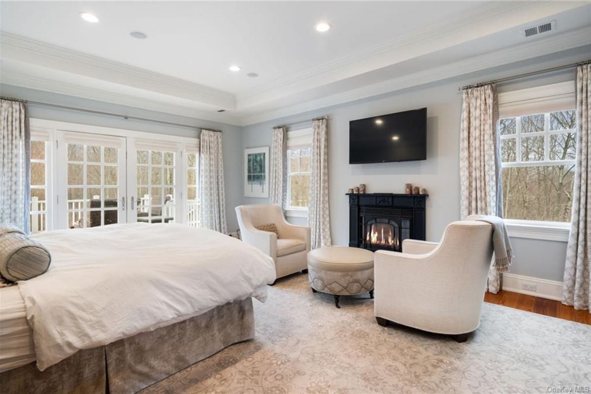 A bedroom with a bed, TV, fireplace, windows, chairs, and a coffee table.
