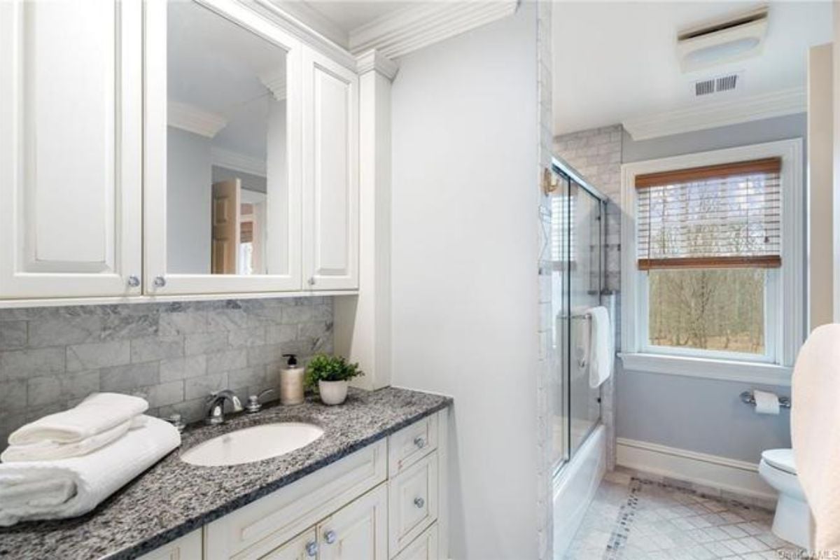 A bathroom with a countertop, sink, mirror, cabinets, toilet, and window.