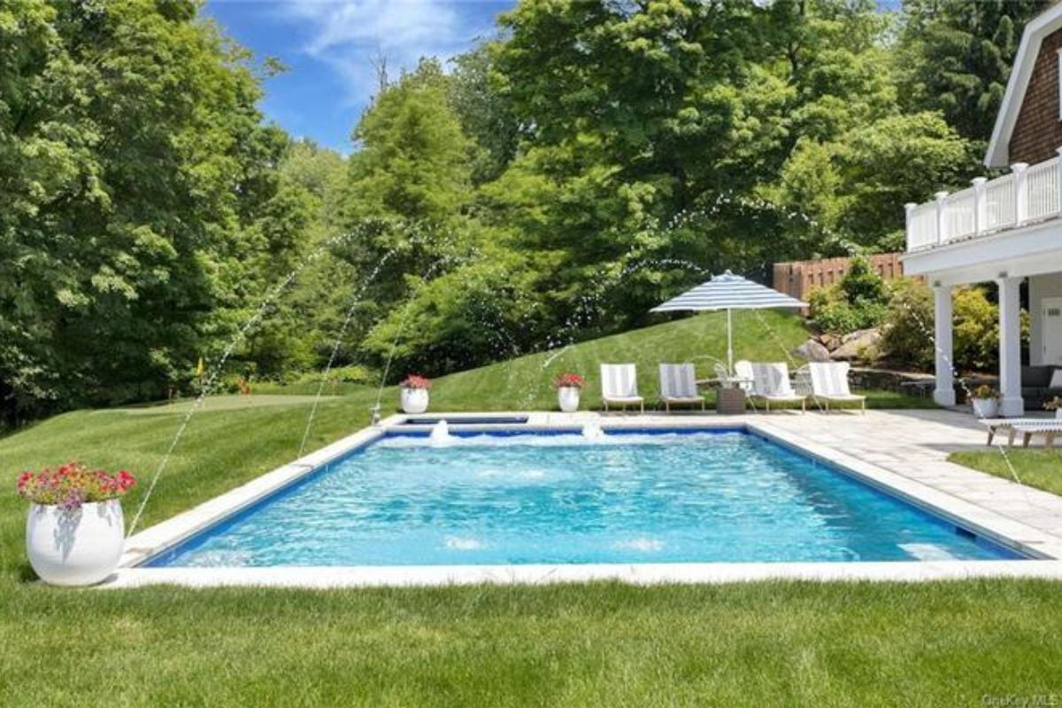 A swimming pool with potted plants and chairs on the poolside, surrounded by grass and trees.