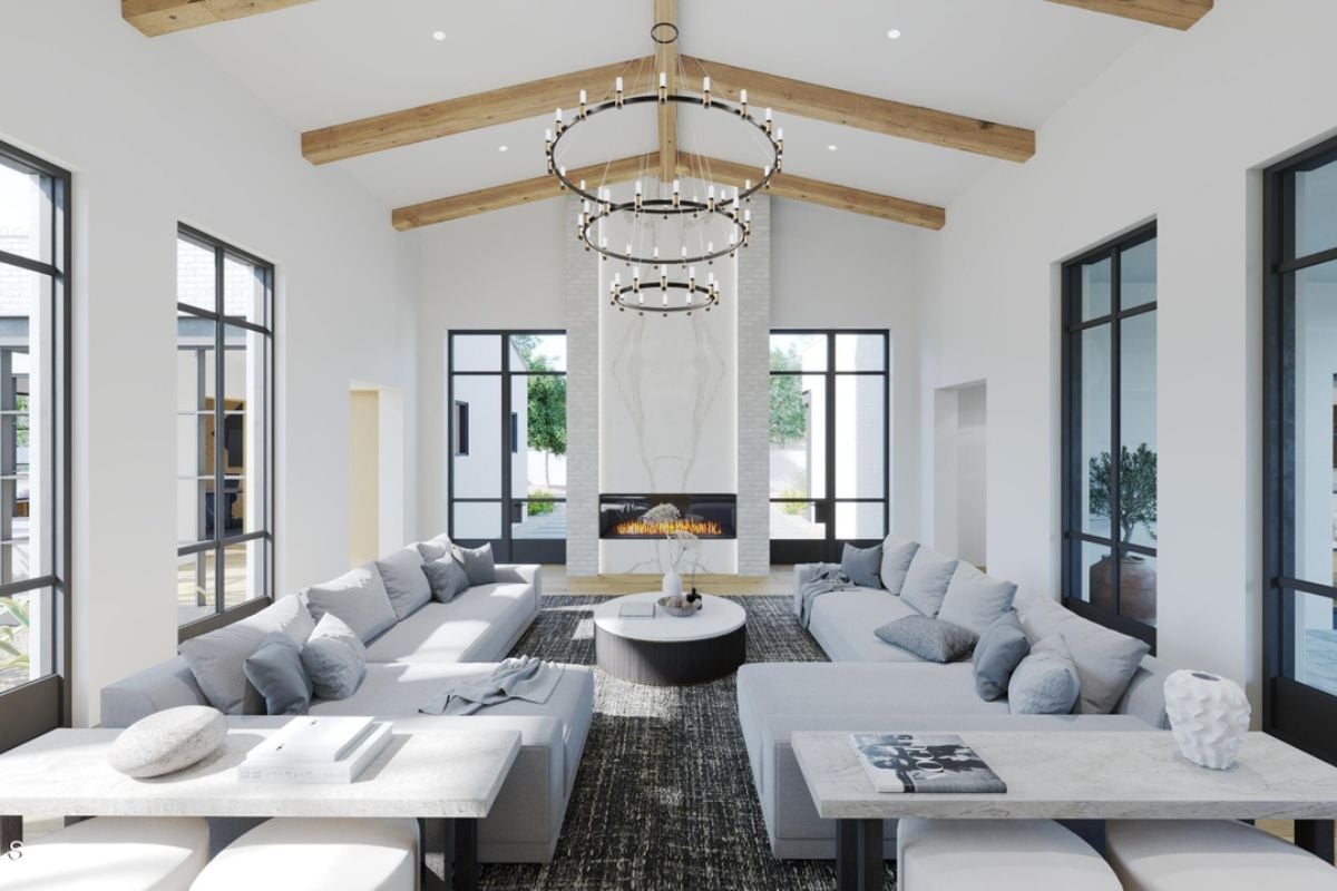 A dining room with a chandelier, exposed beam ceiling, windows, fireplace, white couches, and a round center table.