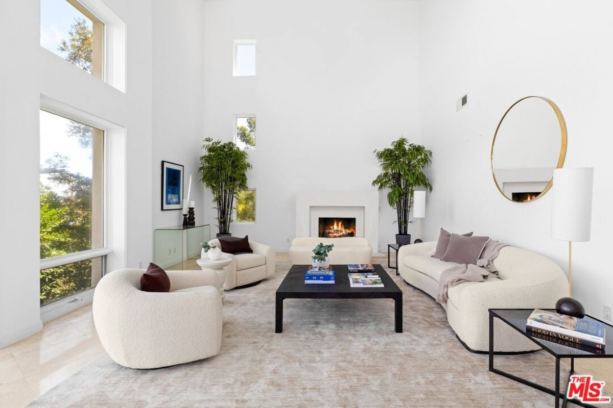 A living room with windows, a fireplace, house plants, a mirror, white couches, and a center table.