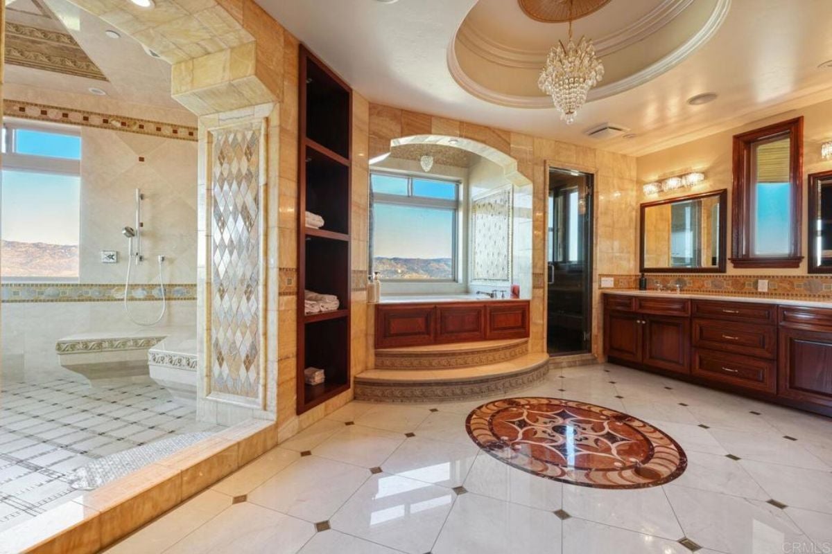 A bathroom with a tiled floor, window, chandelier, countertop, cabinets, mirrors, bathtub, and shower room.