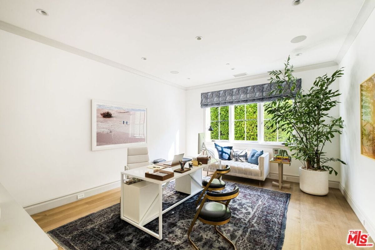 The home office is adorned with house plants, a window, paintings, a couch, a rug, and a desk with chairs.