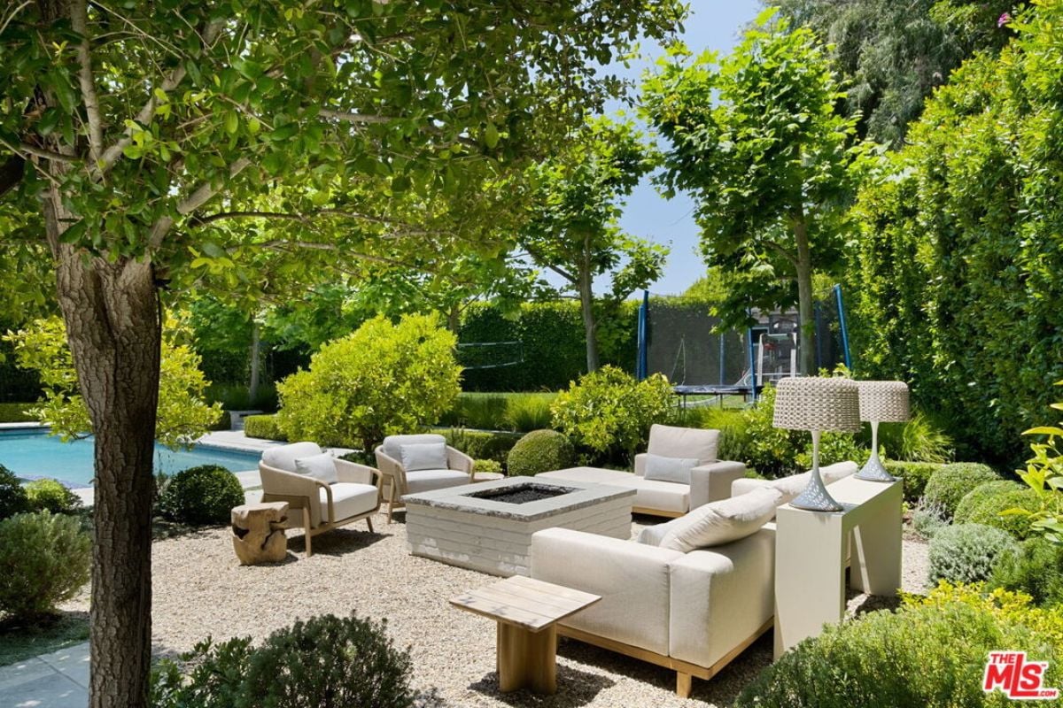The patio, surrounded by plants and trees, features chairs, a couch, and a firepit.