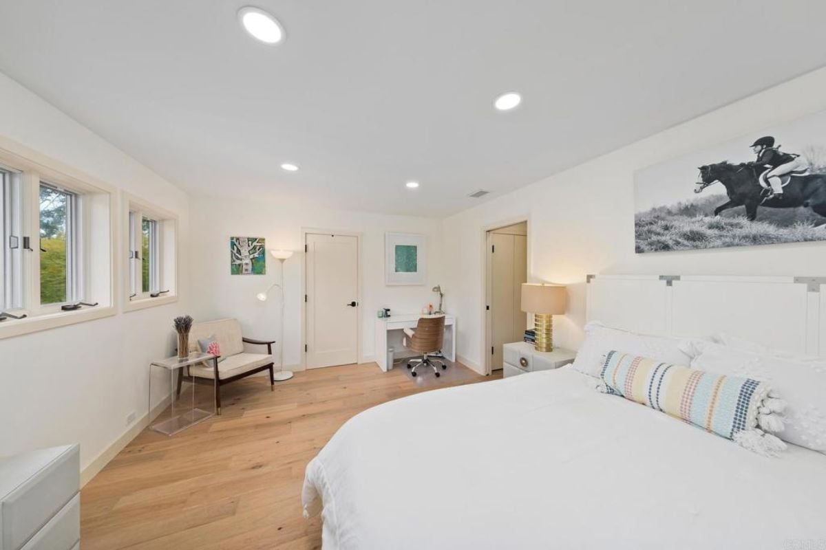 A bedroom with a bed, painting, windows, nightstand, chair, and a desk with chairs.