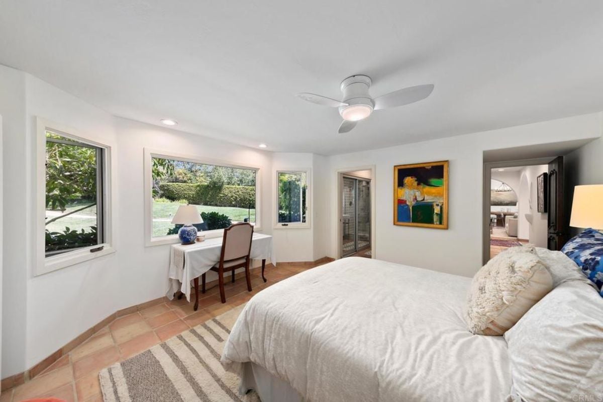 A bedroom with a bed, windows, a ceiling fan, a painting, and a desk with a chair.