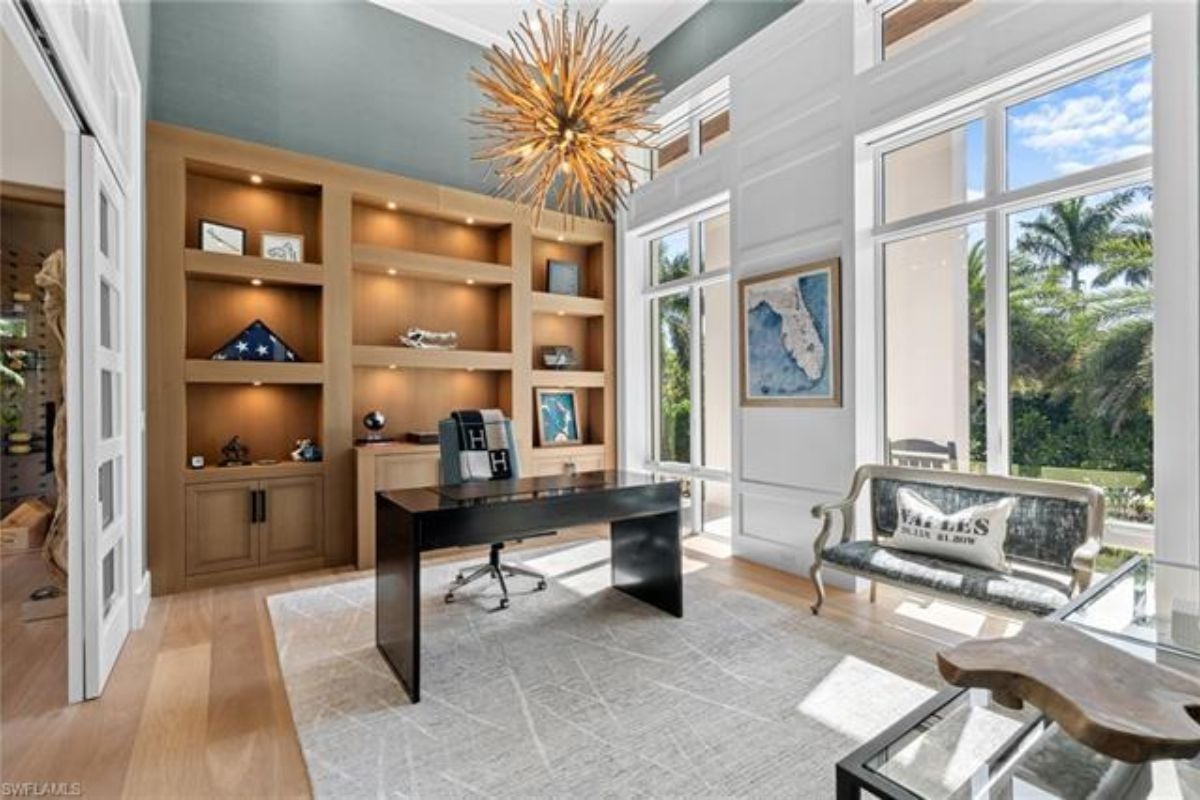 A home office with windows, a chandelier, shelves, a couch, and a desk with a chair.