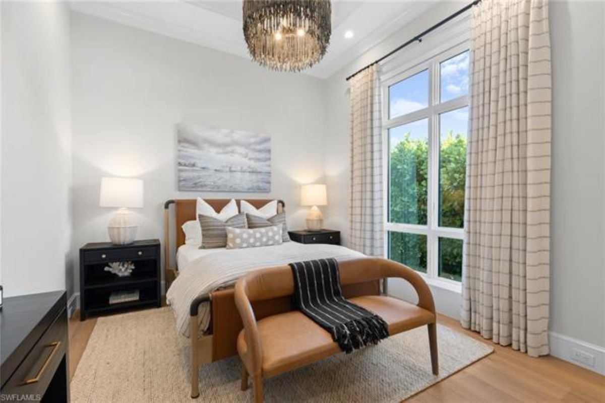 A bedroom with nightstands, a painting, a chandelier, a window, and a bench.