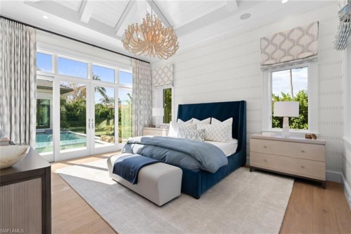 A bedroom with a bed, a chandelier, cabinets, a rug, and windows.