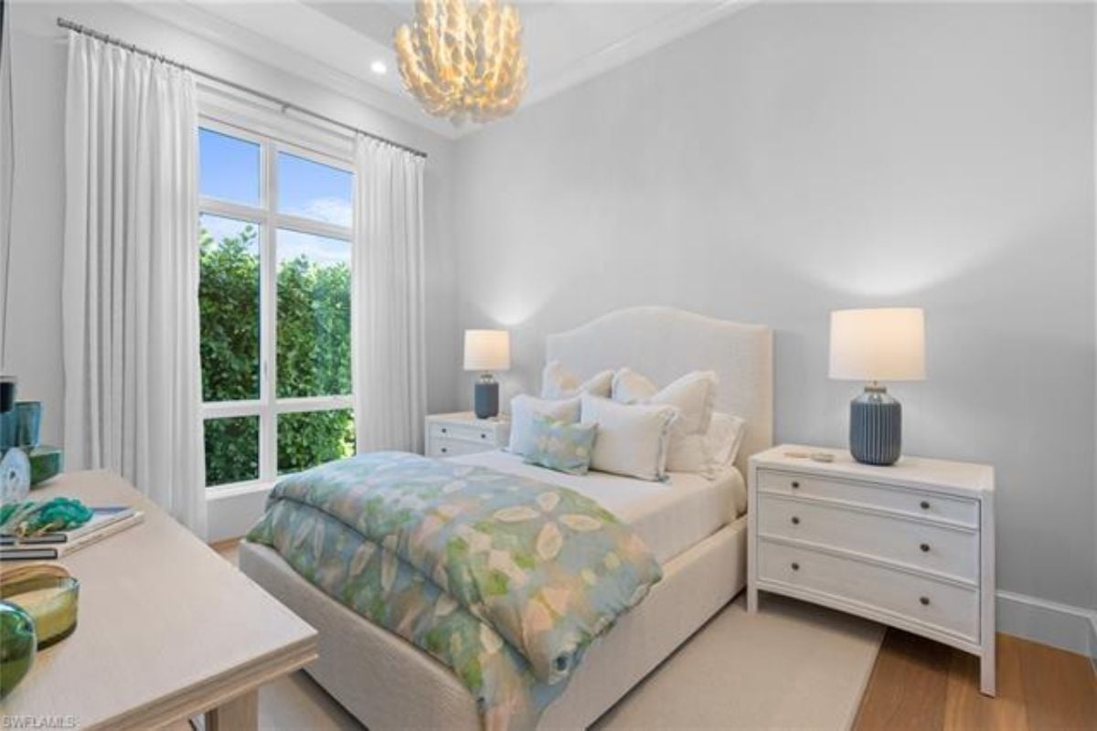 A bedroom with a bed, a chandelier, nightstands, a rug, desk and windows.