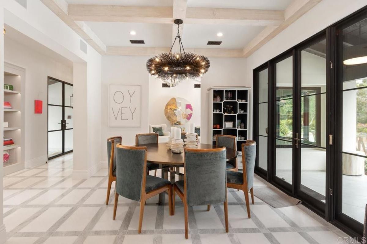 A dining room with an exposed beam ceiling, chandelier, shelves, and a round table surrounded by chairs.