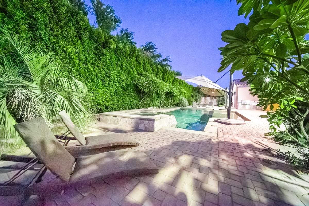 A swimming pool with a jacuzzi, umbrellas, and chairs surrounded by plants.