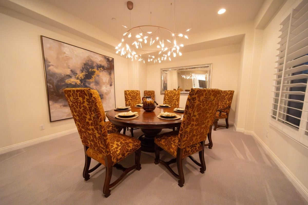 A dining room with a painting, chandelier, and a round dining table surrounded by chairs.