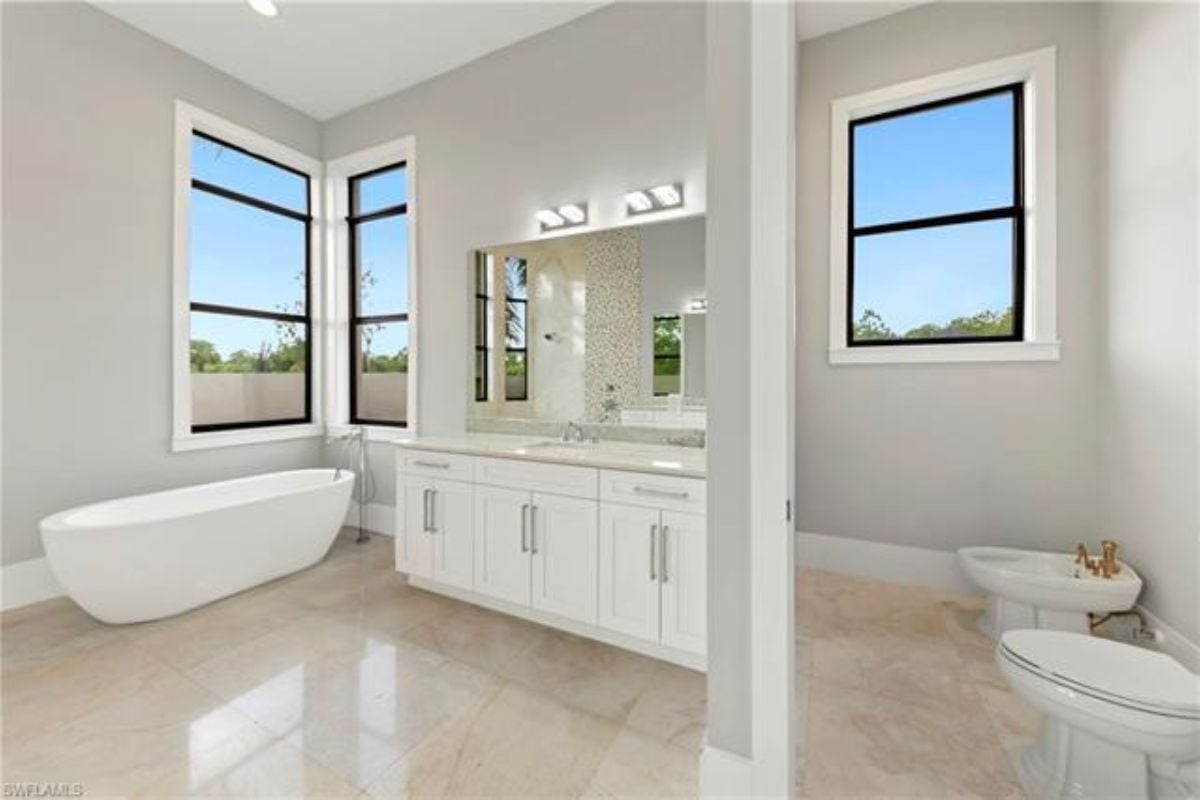 A bathroom with windows, a bathtub, a vanity, a mirror, a cabinet, and a toilet.