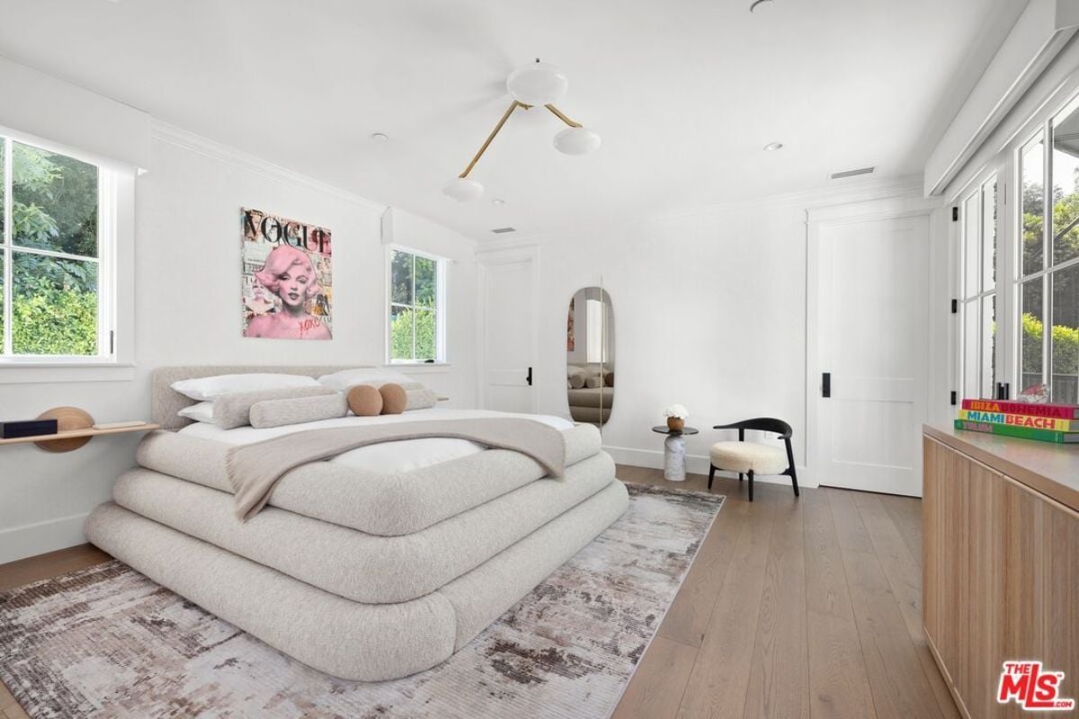 A bedroom with a bed, windows, painting, rug, chair, and coffee table.
