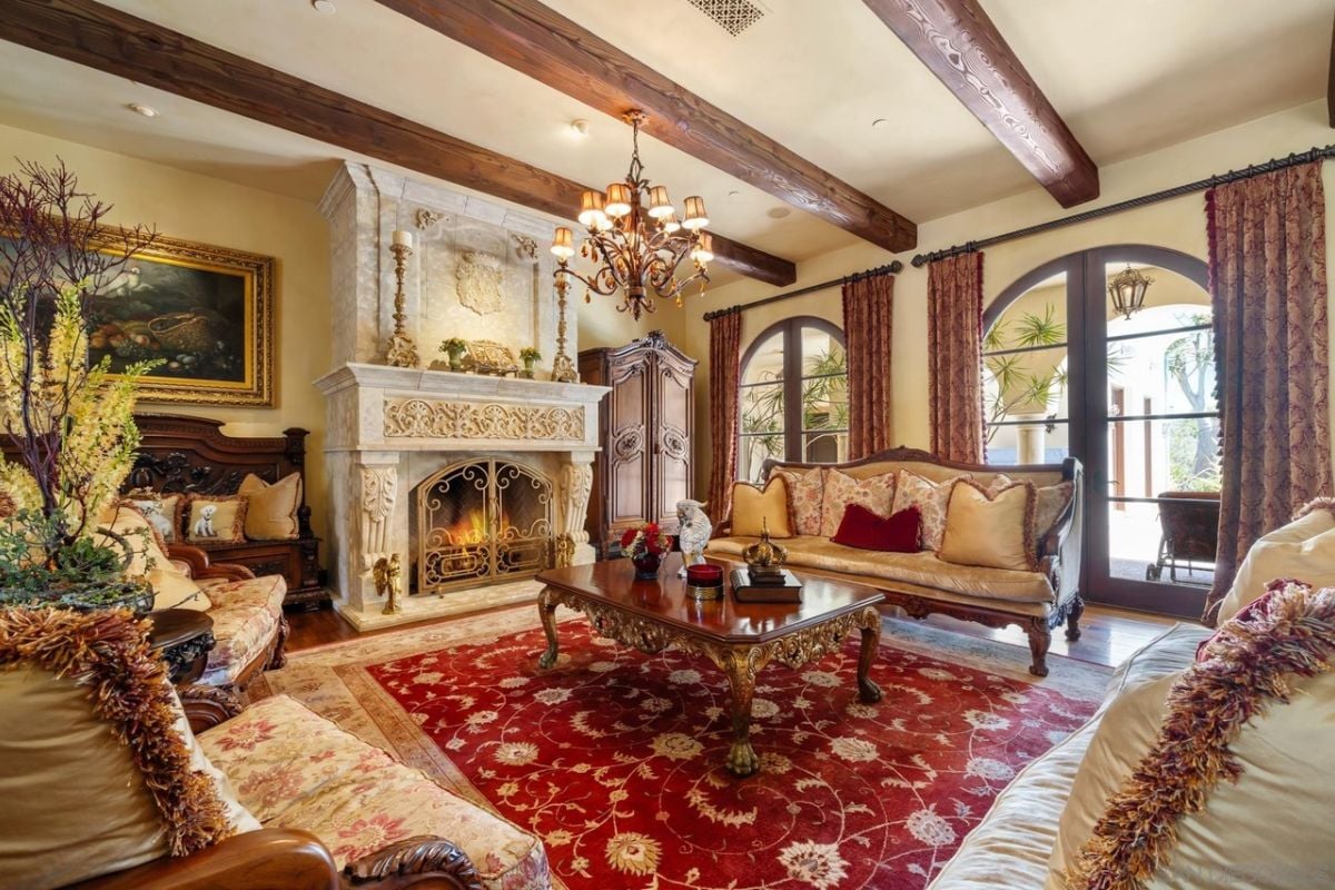 A living room with an exposed beam ceiling, fireplace, painting, windows, rug, couches with pillows, and plants.