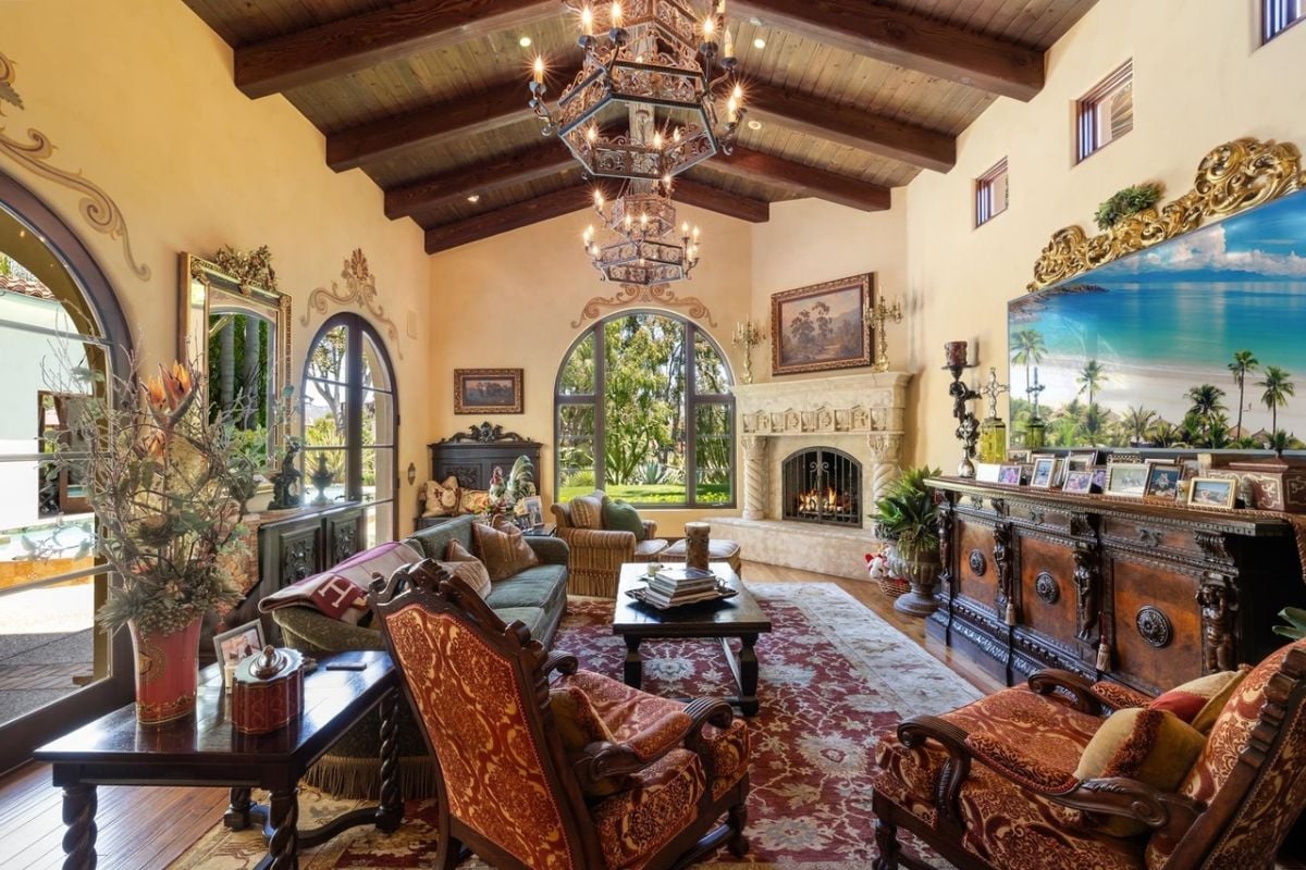 A living room with an exposed beam ceiling, chandeliers, painting, windows, rug, TV, center table, couches with pillows, and plants.