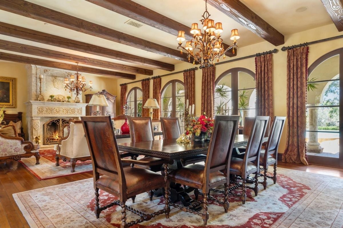 A dining room with an exposed beam ceiling, chandelier, windows, and a dining table surrounded by chairs.