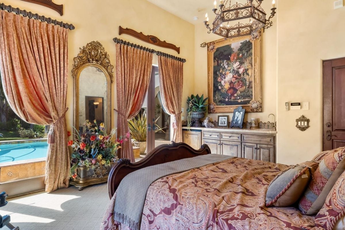 A bedroom with a bed, mirror, windows, painting, cabinet, and plants.