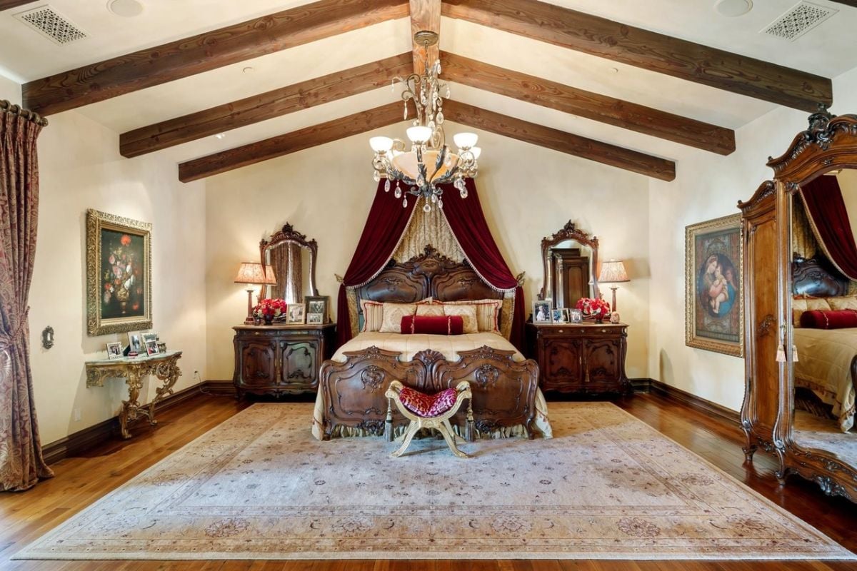 A bedroom with a bed, exposed beam ceiling, chandelier, paintings, cabinets, and mirrors.