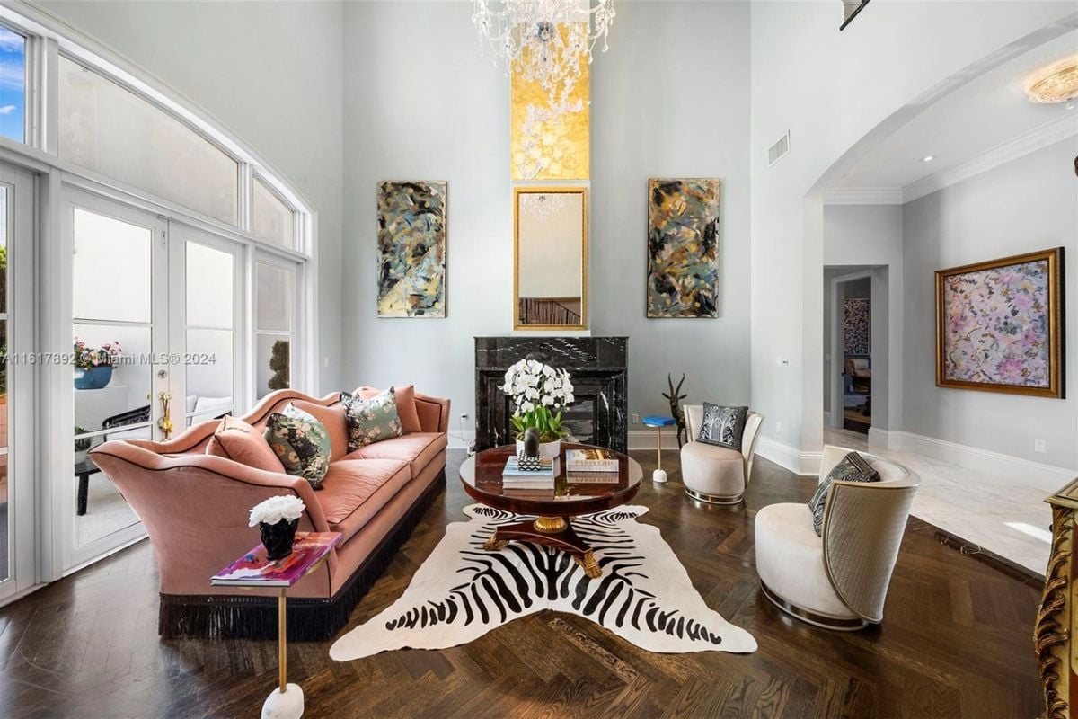 A living room adorned with a chandelier, fireplace, rug, paintings, couch, chairs, and a center table.