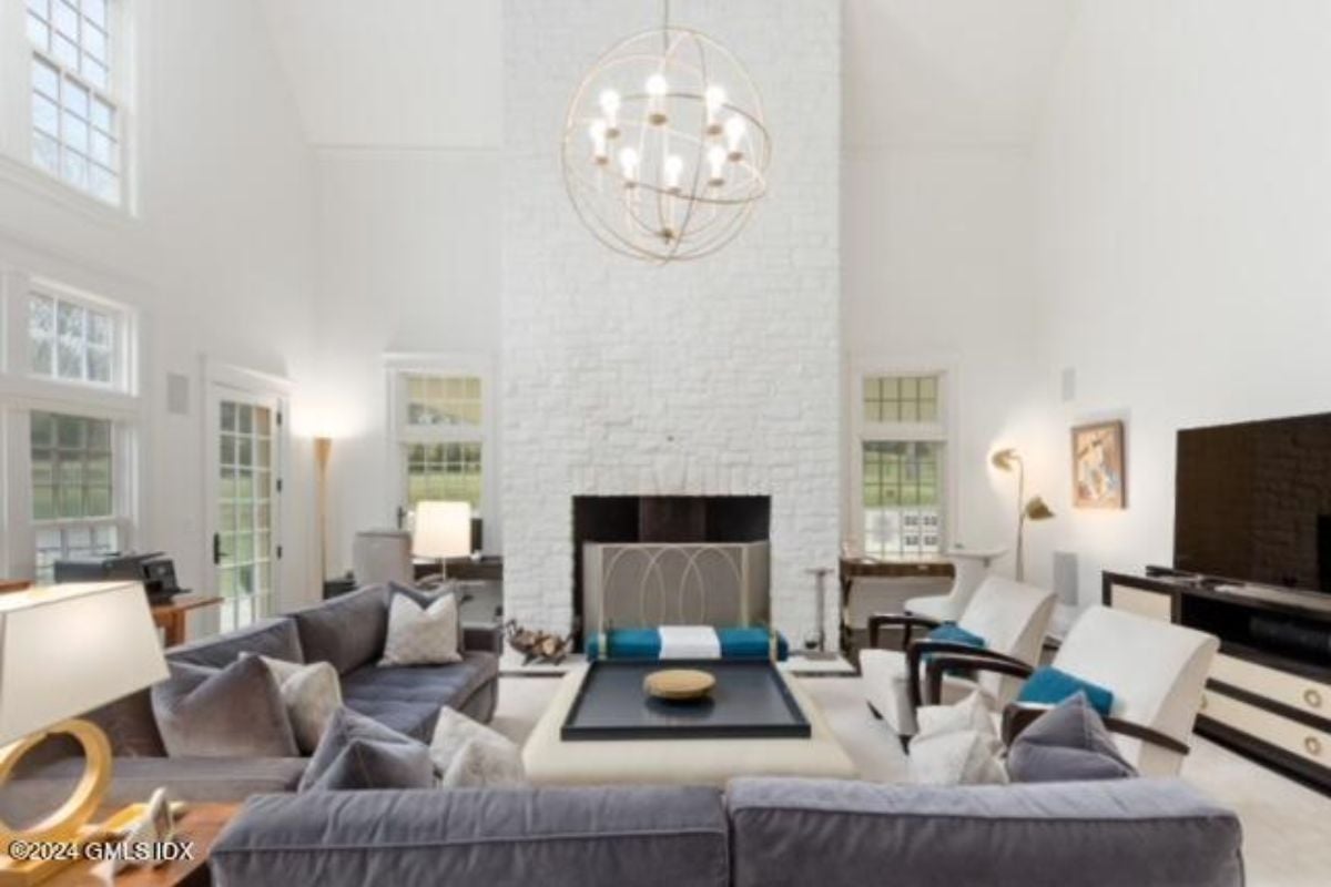 A living room with a high ceiling, chandelier, windows, fireplace, couches, chairs, and a center table.
